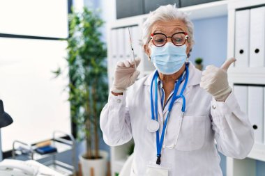 Gri saçlı, doktor üniforması giyen, elinde şırınga tutan ve ağzı açık gülümseyen yaşlı bir kadın. 