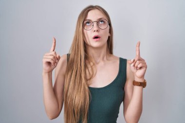 Young caucasian woman standing over white background amazed and surprised looking up and pointing with fingers and raised arms. 