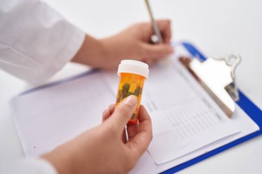 Young beautiful hispanic woman pharmacist holding pills bottle writing on document at pharmacy