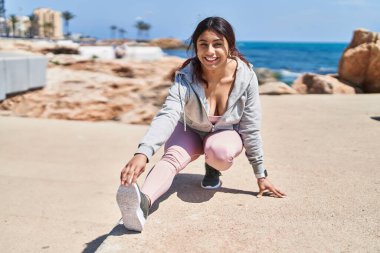 Young hispanic woman stretching legs training at seaside