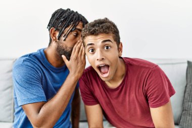 Two men friends whispering secret sitting on sofa at home