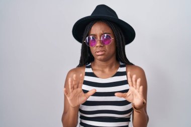 Young african american with braids wearing hat and sunglasses moving away hands palms showing refusal and denial with afraid and disgusting expression. stop and forbidden. 