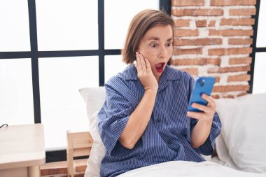 Orta yaşlı bir kadın yatak odasında akıllı telefon kullanıyor.