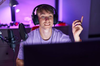Caucasian blond man playing video games smiling happy pointing with hand and finger to the side 