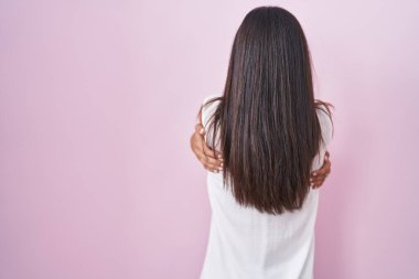 Brunette young woman standing over pink background wearing glasses hugging oneself happy and positive from backwards. self love and self care 