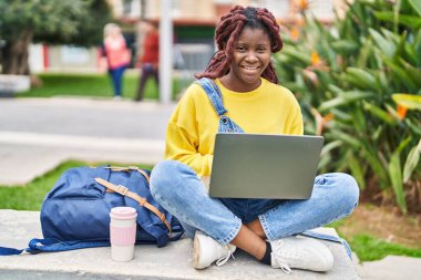 African american woman student using laptop sitting on bench at campus park