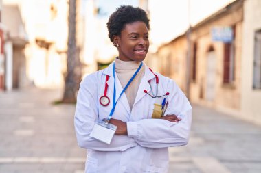 Doktor üniforması giyen Afrikalı Amerikalı kadın caddede kollarını kavuşturmuş duruyor.