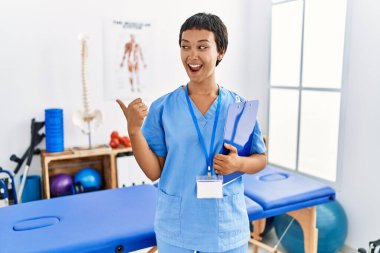 Young hispanic woman with short hair working at pain recovery clinic smiling with happy face looking and pointing to the side with thumb up. 