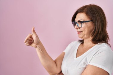 Middle age hispanic woman standing over pink background looking proud, smiling doing thumbs up gesture to the side 