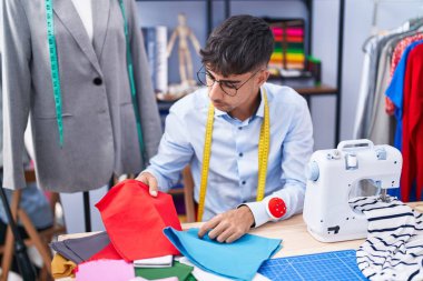 Young hispanic man tailor holding cloths at tailor shop