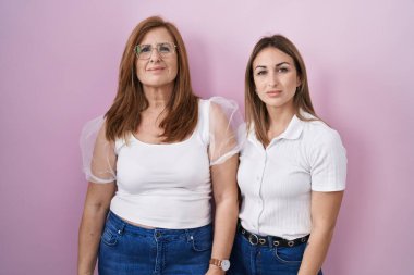 Hispanic mother and daughter wearing casual white t shirt over pink background relaxed with serious expression on face. simple and natural looking at the camera. 