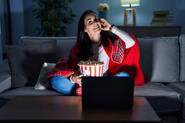 Hispanic woman eating popcorn watching a movie on the sofa smiling confident touching hair with hand up gesture, posing attractive and fashionable 