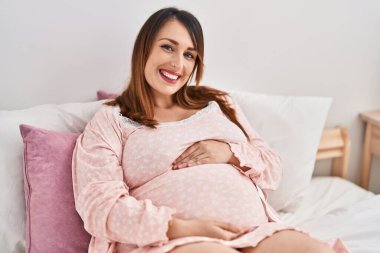 Young pregnant woman touching belly sitting on bed at bedroom