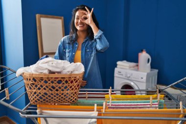 Young asian woman hanging clothes at clothesline doing ok gesture with hand smiling, eye looking through fingers with happy face. 