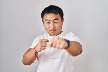 Young chinese man standing over white background punching fist to fight, aggressive and angry attack, threat and violence 