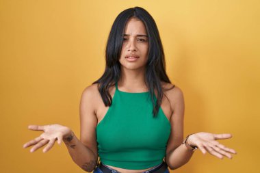 Brunette woman standing over yellow background clueless and confused with open arms, no idea concept. 