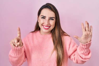 Pembe arka planda duran genç İspanyol kadın altı numaralı parmaklarını gösterip işaret ederken kendinden emin ve mutlu gülümsüyor.. 
