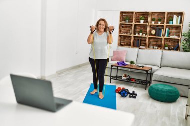 Orta yaşlı beyaz kadın evde elastik bant kullanarak dizüstü bilgisayar eğitimi alıyor.