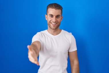 Young caucasian man standing over blue background smiling friendly offering handshake as greeting and welcoming. successful business. 