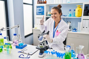 Young hispanic woman working at scientist laboratory surprised with an idea or question pointing finger with happy face, number one 
