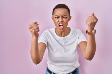 Güzel Afro-Amerikalı kadın pembe arka planda kızgın ve kızgın duruyor öfkeyle bağırırken kızgın ve öfkeli yumruklar sallıyor. öfke ve agresif konsept. 