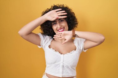 Sarı arka planda duran kıvırcık saçlı genç esmer kadın. Gülümseyen, gülümseyen ve elleri yüzünü gösteren bir yuhalama oyunu oynayan. Şaşırdım ve çıktım. 
