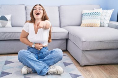 Oturma odasında yerde oturan beyaz bir kadın sana gülüyor, kamerayı işaret ediyor, vücudunu gösteriyor, utanç ifadesi var. 