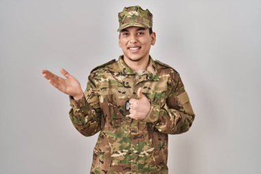 Young arab man wearing camouflage army uniform showing palm hand and doing ok gesture with thumbs up, smiling happy and cheerful 