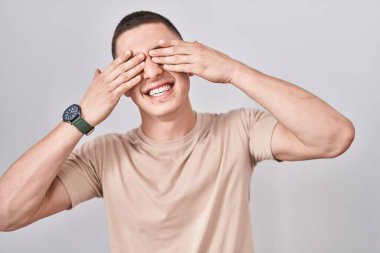 İzole edilmiş arka planda duran genç adam elleri gülücüklü ve komik bir şekilde gözlerini kapatıyor. kör kavram. 