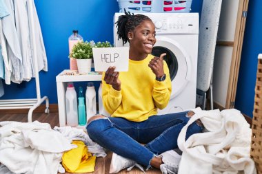 Beautiful black woman doing laundry asking for help pointing thumb up to the side smiling happy with open mouth 