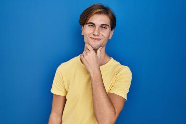 Young man standing over blue background looking confident at the camera smiling with crossed arms and hand raised on chin. thinking positive. 