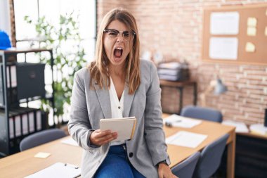 Young hispanic woman working at the office wearing glasses angry and mad screaming frustrated and furious, shouting with anger. rage and aggressive concept. 