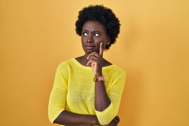 African young woman standing over yellow studio thinking worried about a question, concerned and nervous with hand on chin 