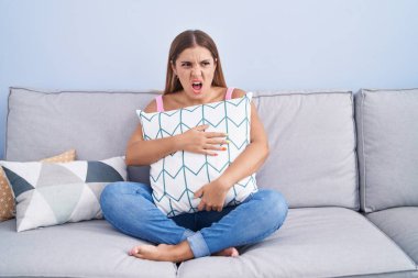 Young blonde woman hugging pillow sitting on the sofa angry and mad screaming frustrated and furious, shouting with anger. rage and aggressive concept. 
