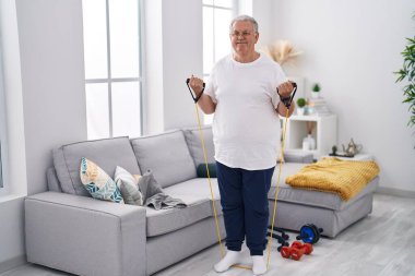 Orta yaşlı, gri saçlı, kendine güvenen, evde elastik bant eğitimi alan bir adam.