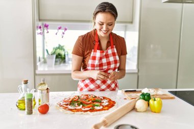 Young beautiful hispanic woman smiling confident putting onion on pizza at the kitchen