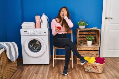 Young hispanic woman sitting waiting for laundry using smartphone yawning tired covering half face, eye and mouth with hand. face hurts in pain. 