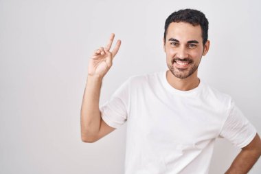 Handsome hispanic man standing over white background smiling looking to the camera showing fingers doing victory sign. number two. 