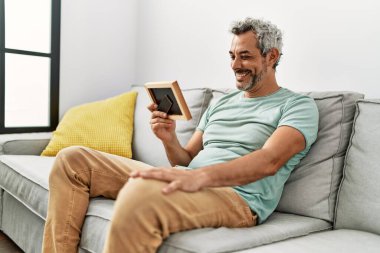 Orta yaşlı, kır saçlı, evdeki kanepede oturan bir adam.