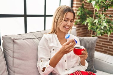 Young hispanic woman drinking coffee pouring saccharin at home