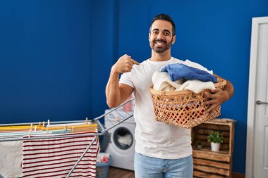 Sakallı, çamaşır sepetini tutarak gülümseyen mutlu ve gururlu birini işaret eden genç bir İspanyol. 