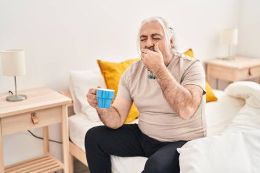Middle age grey-haired man sitting on bed yawning and drinking coffee at bedroom