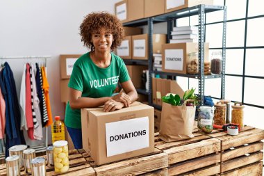 Young african american woman wearing volunteer uniform leaning on donations box at charity center