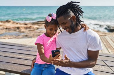 Father and daughter using smartphone hugging each other at seaside