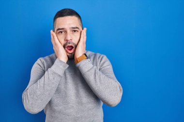 Mavi arka plan üzerinde duran İspanyol adam korkmuş ve şok olmuş, şaşırmış ve şaşırmış bir yüz ifadesi var. 