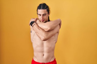 Hispanic man with long hair standing shirtless over yellow background hugging oneself happy and positive, smiling confident. self love and self care 