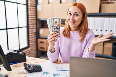 Küçük işletme şirketinde çalışan genç bir kadın elinde para tutarak başarıyı mutlu bir gülümsemeyle kutluyor ve kazanan el kaldırarak ifade ediyor. 