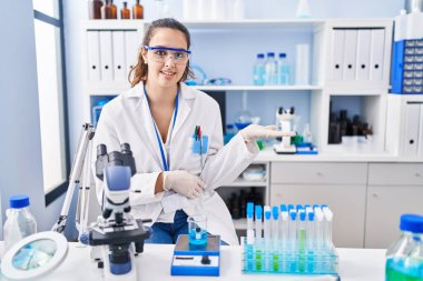 Young hispanic woman working at scientist laboratory smiling cheerful presenting and pointing with palm of hand looking at the camera. 