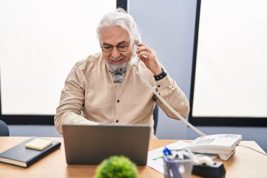 Middle age grey-haired man business worker talking on telephone using laptop at office