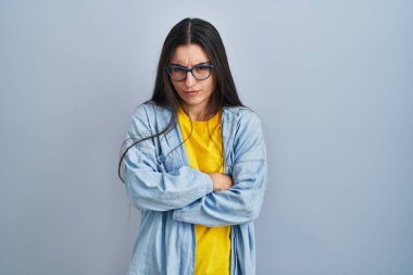 Mavi arka planda duran Latin kökenli genç kadın şüpheci ve gergin, yüzünde çapraz kollu tasvip etmeyen ifade. negatif kişi. 